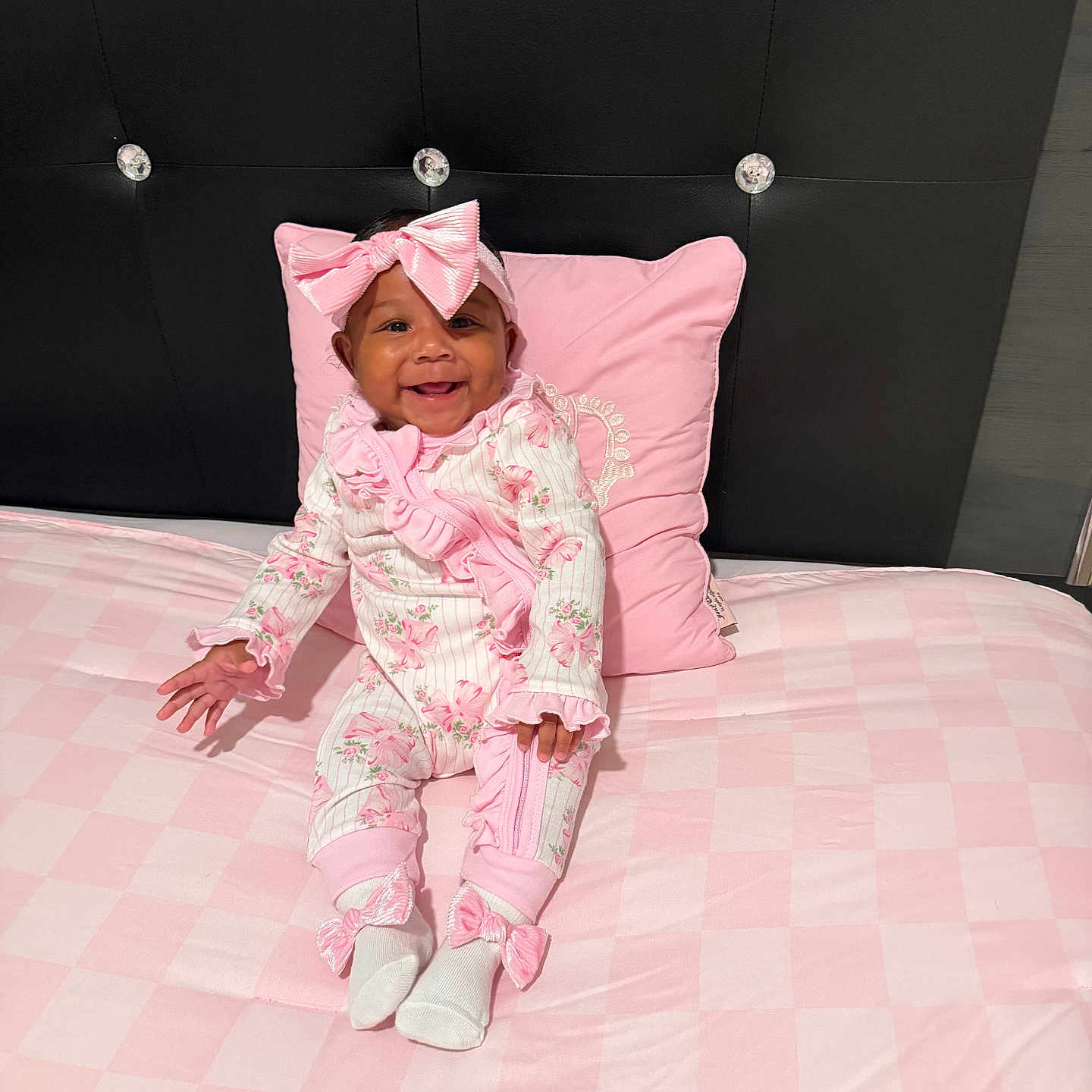 Journi is registered to the contest to win money with this photo: baby, infant, smiling, pink, bow, pajamas, bed, pillow, headboard, checkered, floral, clothing, cute, happy, sitting, indoors, portrait, child, person, soft