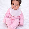 Khaleessi is registered to the contest to win money with this photo: baby, child, infant, smile, pink_onesie, bib, curly_hair, sitting, soft_blanket, cute, portrait, indoors, happy, young_child, cozy, hands_covered, feet_covered, face, person, toddler