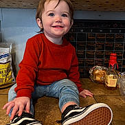 Phenix is registered to the contest to win money with this photo: toddler, child, smiling, orange_sweater, jeans, sneakers, kitchen, countertop, floor_tiles, chalkboard_calendar, bag_of_sugar, bottle, bread, happy, blue_eyes, casual_clothing, indoor, person, cute, young_child
