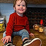 toddler, child, smiling, orange_sweater, jeans, sneakers, kitchen, countertop, floor_tiles, chalkboard_calendar, bag_of_sugar, bottle, bread, happy, blue_eyes, casual_clothing, indoor, person, cute, young_child