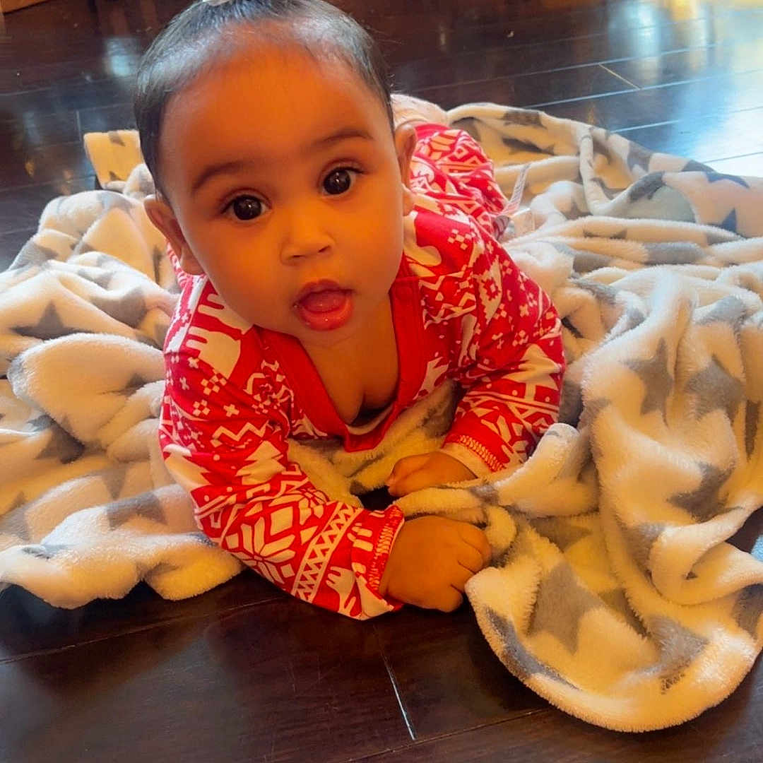 Alani-Rae is registered to the contest to win money with this photo: baby, blanket, child, christmas_tree, cute, decorations, face, festive, floor, holiday, holiday_season, indoors, pajamas, person, presents, smiling, star_pattern, warm_lighting, wooden_floor, young_child