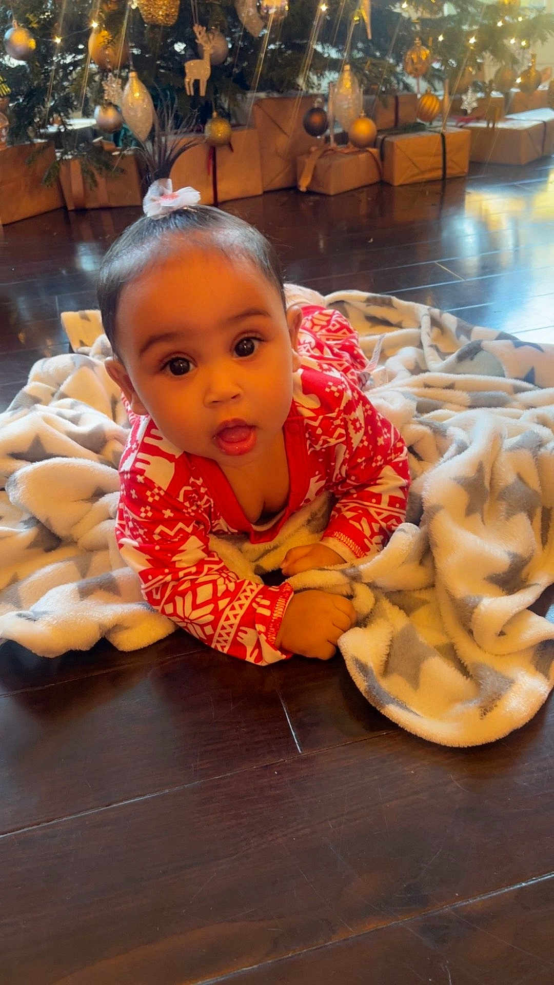 Alani-Rae is registered to the contest to win money with this photo: baby, child, pajamas, blanket, floor, christmas_tree, presents, holiday, indoors, young_child, cute, festive, decorations, star_pattern, wooden_floor, face, smiling, person, warm_lighting, holiday_season