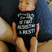 Kaiden is registered to the contest to win money with this photo: baby, newborn, infant, onesie, text_on_clothing, blanket, quilt, legs, tattoo, skin, indoors, resting, cute, person, lying_down, expression, human, clothing, comfort, closeup