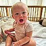 baby, child, lip_gloss, bed, curtains, wooden_bed_frame, pink_clothing, sitting, surprised_expression, indoors, natural_light, toy, blanket, cute, person, face, hair_ponytail, skin, hand, mouth_open