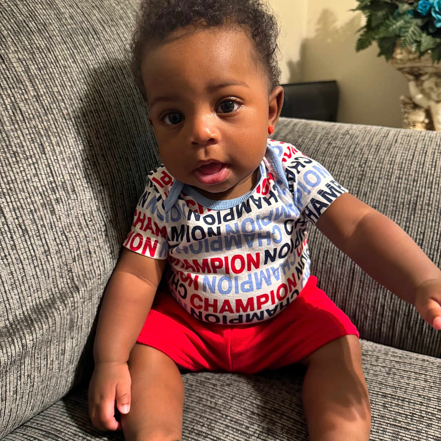 Dakota Haile joined the competition — help win amazing prizes! baby, child, infant, portrait, sitting, couch, indoors, curious, face, smiling, clothing, red_shorts, black_shoes, curly_hair, floral_arrangement, living_room, furniture, closeup, photography, championship_shirt