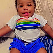 Dakota Haile joined the competition — help win amazing prizes! baby, child, infant, smiling, happy, portrait, face, curly_hair, indoors, bed, bedding, pillow, blanket, striped_pattern, rainbow_shirt, blue_shorts, sitting, lying_down, bedroom, photography