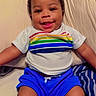 baby, child, infant, smiling, happy, portrait, face, curly_hair, indoors, bed, bedding, pillow, blanket, striped_pattern, rainbow_shirt, blue_shorts, sitting, lying_down, bedroom, photography