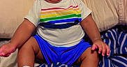 Dakota Haile joined the competition — help win amazing prizes! baby, child, infant, smiling, sitting, portrait, bed, bedroom, striped_bedding, pillow, clothing, rainbow_shirt, blue_shorts, sneakers, curly_hair, indoor, happy, playful, closeup, photography