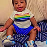 baby, child, infant, smiling, sitting, portrait, bed, bedroom, striped_bedding, pillow, clothing, rainbow_shirt, blue_shorts, sneakers, curly_hair, indoor, happy, playful, closeup, photography