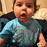 baby, bed, blanket, blue_shirt, casual_clothing, child, curious, curly_hair, cute, facial_expression, hand, indoor, open_mouth, pacifier, person, pillow, portrait, sitting, toddler, young_child
