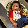 Marcel joined the competition — help win amazing prizes! baby, smiling, curly_hair, infant, red_shirt, navy_pants, baby_seat, patterned_cushion, happy, child, cute, indoor, person, clothing, sitting, young_child, joyful, portrait, dimples, relaxing