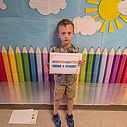 Michael joined the competition — help win amazing prizes! child, boy, sign, kindergarten, colorful_background, crayons, sun, clouds, school, indoor, floor, shoes, shorts, tshirt, person, portrait, standing, expression, paper, education