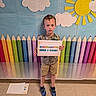 child, boy, sign, kindergarten, colorful_background, crayons, sun, clouds, school, indoor, floor, shoes, shorts, tshirt, person, portrait, standing, expression, paper, education