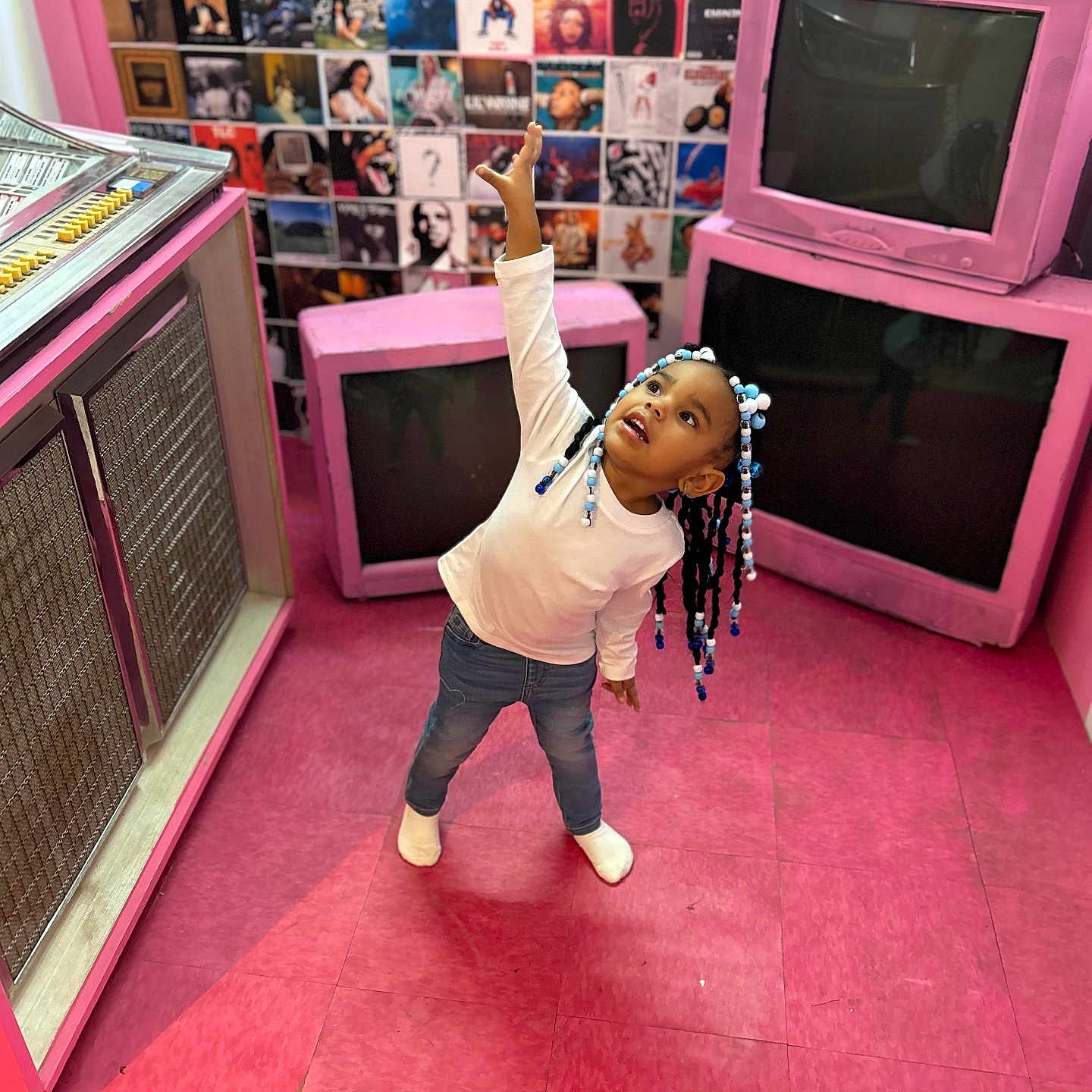 Lamari is registered to the contest to win money with this photo: album_covers, beads, child, colorful, expression, floor, girl, hair, indoor, jeans, pink_floor, playful, pose, retro, room, socks, vintage_tv, wall_collage, white_shirt, young