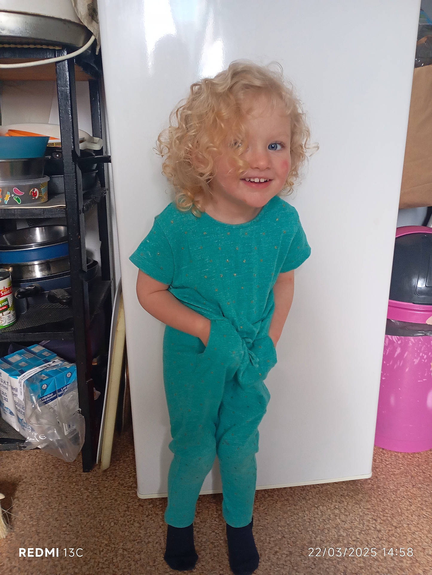 Naila a rejoint le concours — aidez-le/la à gagner de superbes lots ! blonde_hair, blue_eyes, carpet, casual, child, clothing, curly_hair, fridge, hands_in_pockets, happy, household_items, indoor, kitchen, person, portrait, shelf, smiling, standing, teal_outfit, trash_bin