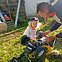 adult, cap, casual_clothing, child, childhood, curly_hair, daylight, fence, garden_tools, grass, hands, lawn, outdoor, person, playing, repair, summer, sunlight, toy_car, vehicle