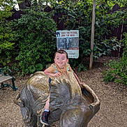Liam is registered to the contest to win money with this photo: child, smile, elephant_statue, bronze, park, outdoor, greenery, bushes, fence, sign, pledge, conservation, happy, playful, sneakers, shorts, tshirt, daylight, nature, woodchips
