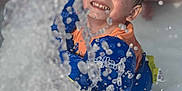 Liam is registered to the contest to win money with this photo: child, smiling, water, splash, life_jacket, play, happy, outdoor, summer, pool, water_sport, fun, person, blue, orange, sitting, wet, joy, closeup, face