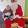 baby, santa_claus, child, holiday, costume, indoor, book, person, red_clothing, beard, gloves, boots, chair, floor, wall, clock, toy, decorations, striped_clothing, smile