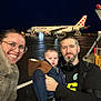 airplane, airport, baby, beard, coat, family, glasses, happy, headphones, jacket, luggage_cart, man, night, outdoor, people, smile, stairs, tarmac, travel, woman