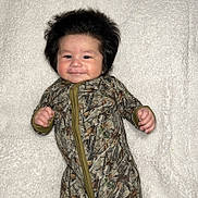 Joseph joined the competition — help win amazing prizes! baby, infant, face, hair, smile, camouflage, onesie, blanket, lying_down, cheeks, cute, child, person, soft_texture, patterned_clothing, indoors, portrait, happy, skin, young