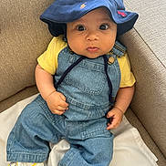 Dayan participe au concours pour gagner de l'argent avec cette photo : baby, bucket_hat, casual, child, clothing, comfortable, couch, curious, cute, denim_overalls, fabric, footwear, indoor, infant, portrait, seated, sneakers, white, wide_eyes, yellow