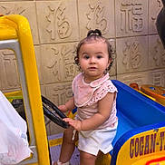 Aleyna participe au concours pour gagner de l'argent avec cette photo : blue_seat, child, curly_hair, cute, decorative_wall, expression, footwear, indoor, kiddie_ride, pink_clothing, playful, sandals, sitting, small_person, steering_wheel, summer_clothes, toddler, toy_car, wall_art, yellow_car