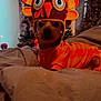 dog, pet, costume, turkey_hat, orange_outfit, bed, blanket, christmas_tree, holiday_decor, indoor, festive, cozy, cute, animal, warm_lighting, decoration, holiday_season, soft_texture, small_dog, portrait