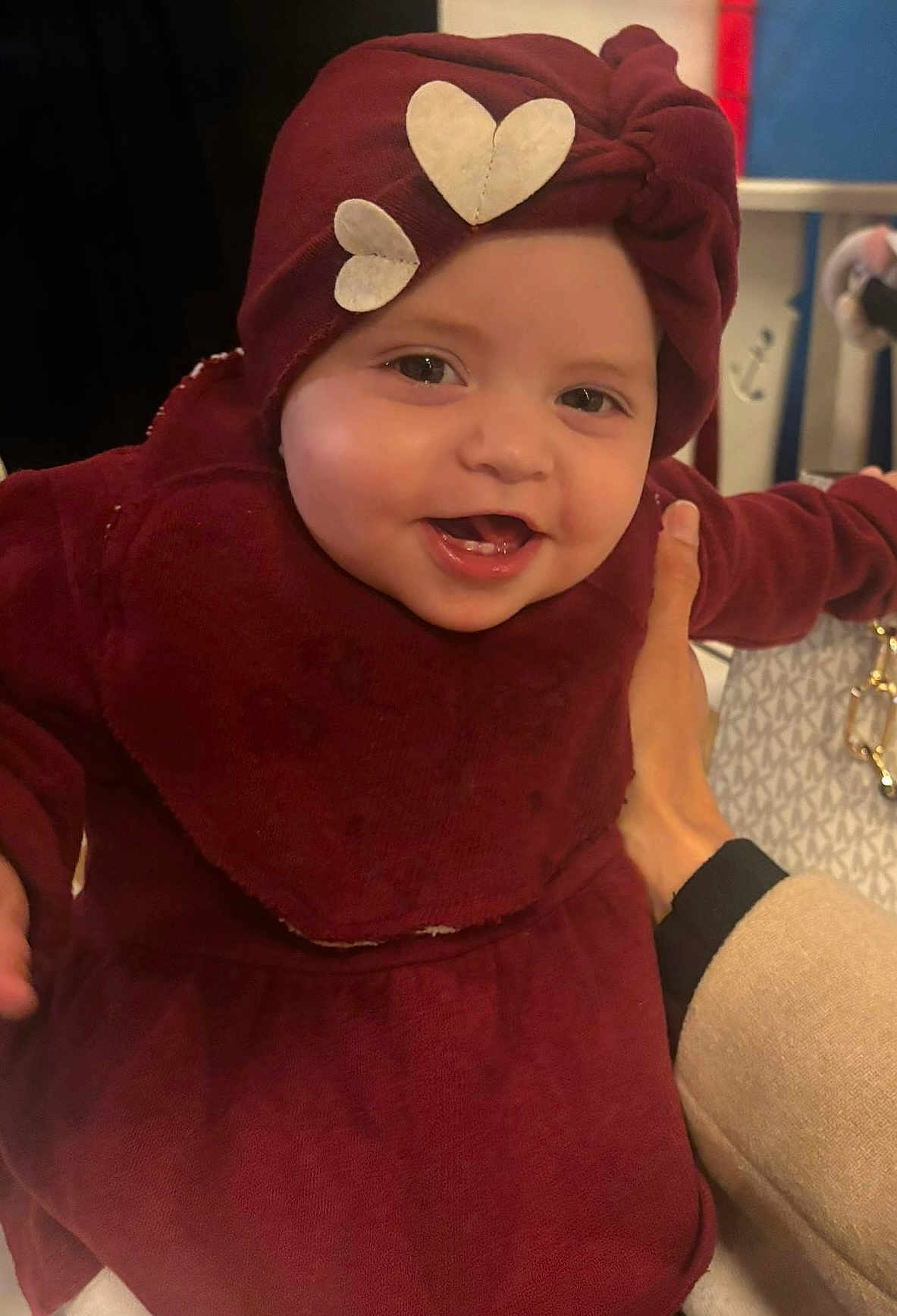 Louna a rejoint le concours — aidez-le/la à gagner de superbes lots ! baby, child, smiling, maroon_clothing, headwrap, heart_shapes, hand, indoor, person, face, happy, holding, fabric, patterned_bag, adult_hand, cute, portrait, closeup, infant, warm_clothing