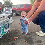 Rhaenyra is registered to the contest to win money with this photo: baby, child, man, overalls, denim, wrench, car, license_plate, street, vehicle, red_truck, smile, happy, barefoot, person, outdoor, asphalt, tool, supporting_hand, daylight