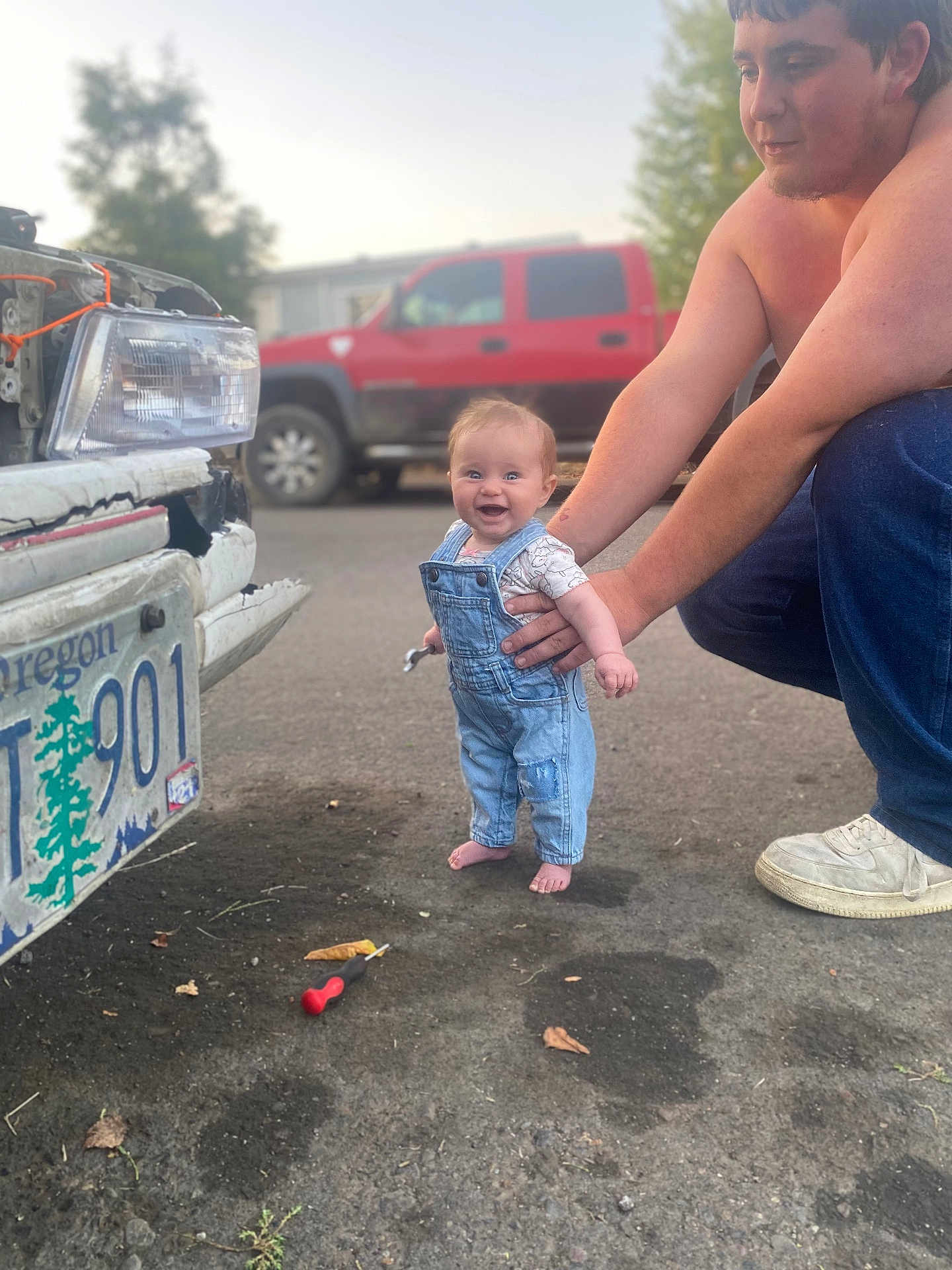 Rhaenyra is registered to the contest to win money with this photo: baby, child, man, overalls, denim, wrench, car, license_plate, street, vehicle, red_truck, smile, happy, barefoot, person, outdoor, asphalt, tool, supporting_hand, daylight