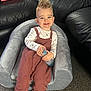 toddler, child, smiling, bottle, chair, plush_chair, knitted_overalls, long_sleeve_shirt, blue_eyes, spiky_hair, carpet, leather_couch, indoor, red_box, nintendo_box, footwear, happy, comfortable, casual_clothing, home_interior