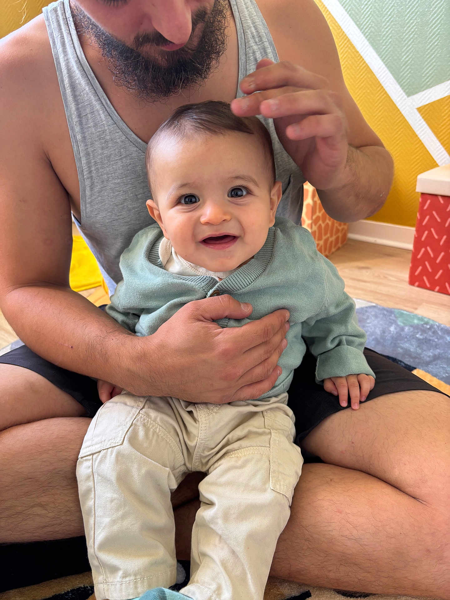 Leeroy a rejoint le concours — aidez-le/la à gagner de superbes lots ! baby, man, smile, indoor, child, holding, sitting, clothing, pants, tank_top, beard, floor, carpet, hand, face, person, room, wall, love, family