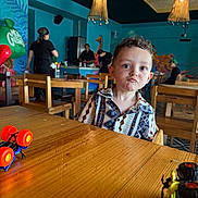 Mateo joined the competition — help win amazing prizes! child, toddler, boy, portrait, pouting, restaurant, cafe, indoor, table, wooden_table, chairs, toy_cars, colorful, mural, pendant_lights, seated, patterned_shirt, curly_hair, people, dining_area