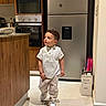 child, boy, toddler, portrait, standing, walking, indoors, kitchen, refrigerator, oven, microwave, wooden_cabinets, white_shirt, striped_pants, sneakers, fashion, curly_hair, facial_expression, home, wine_box