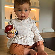 Ilyana participe au concours pour gagner de l'argent avec cette photo : toddler, child, toy, table, indoor, blue_eyes, hair_clip, white_blouse, brown_pants, barefoot, wood_floor, modern_interior, sitting, curious, portrait, person, cute, lighting, decor, furniture