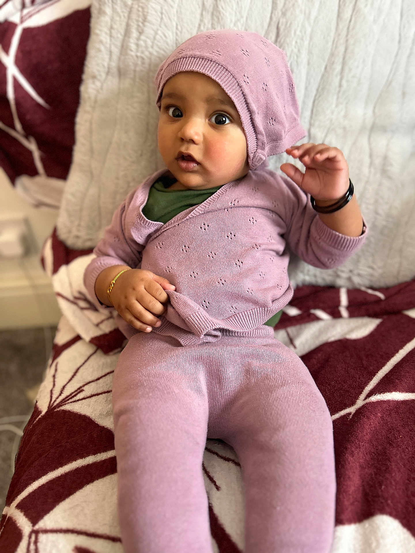 Jordan is registered to the contest to win money with this photo: baby, child, infant, purple_clothing, hat, bracelet, sitting, blanket, pattern, indoor, cute, portrait, knitwear, expression, person, young_child, cozy, soft_texture, curious, wide_eyes