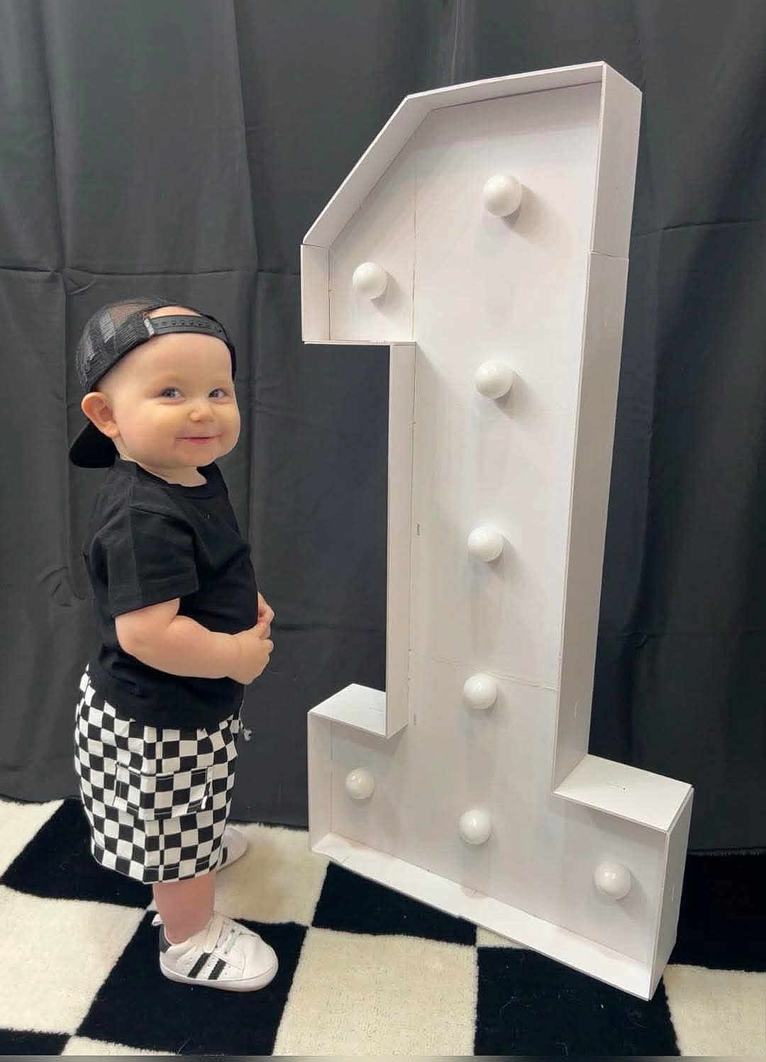 Drew is registered to the contest to win money with this photo: toddler, child, boy, black_shirt, checkered_shorts, white_shoes, black_cap, smiling, standing, number_one, decor, birthday, indoor, floor, patterned_floor, black_background, white_lights, cute, portrait, celebration