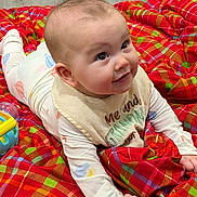 Ember joined the competition — help win amazing prizes! baby, infant, child, smiling, lying_down, bib, blanket, plaid, colorful, toy, floor, indoors, cute, face, hands, clothing, happy, expression, person, portrait