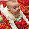 baby, infant, child, smiling, lying_down, bib, blanket, plaid, colorful, toy, floor, indoors, cute, face, hands, clothing, happy, expression, person, portrait