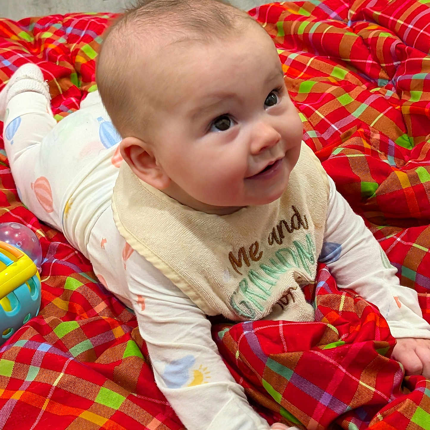 Ember joined the competition — help win amazing prizes! baby, infant, child, smiling, lying_down, bib, blanket, plaid, colorful, toy, floor, indoors, cute, face, hands, clothing, happy, expression, person, portrait