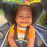 Ahsai is registered to the contest to win money with this photo: toddler, child, smiling, orange_shirt, stroller, seatbelt, food_mess, outdoor, playground, grass, greenery, happy, baby, face, person, cute, curly_hair, sunlight, play_equipment, seat