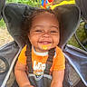 toddler, child, smiling, orange_shirt, stroller, seatbelt, food_mess, outdoor, playground, grass, greenery, happy, baby, face, person, cute, curly_hair, sunlight, play_equipment, seat