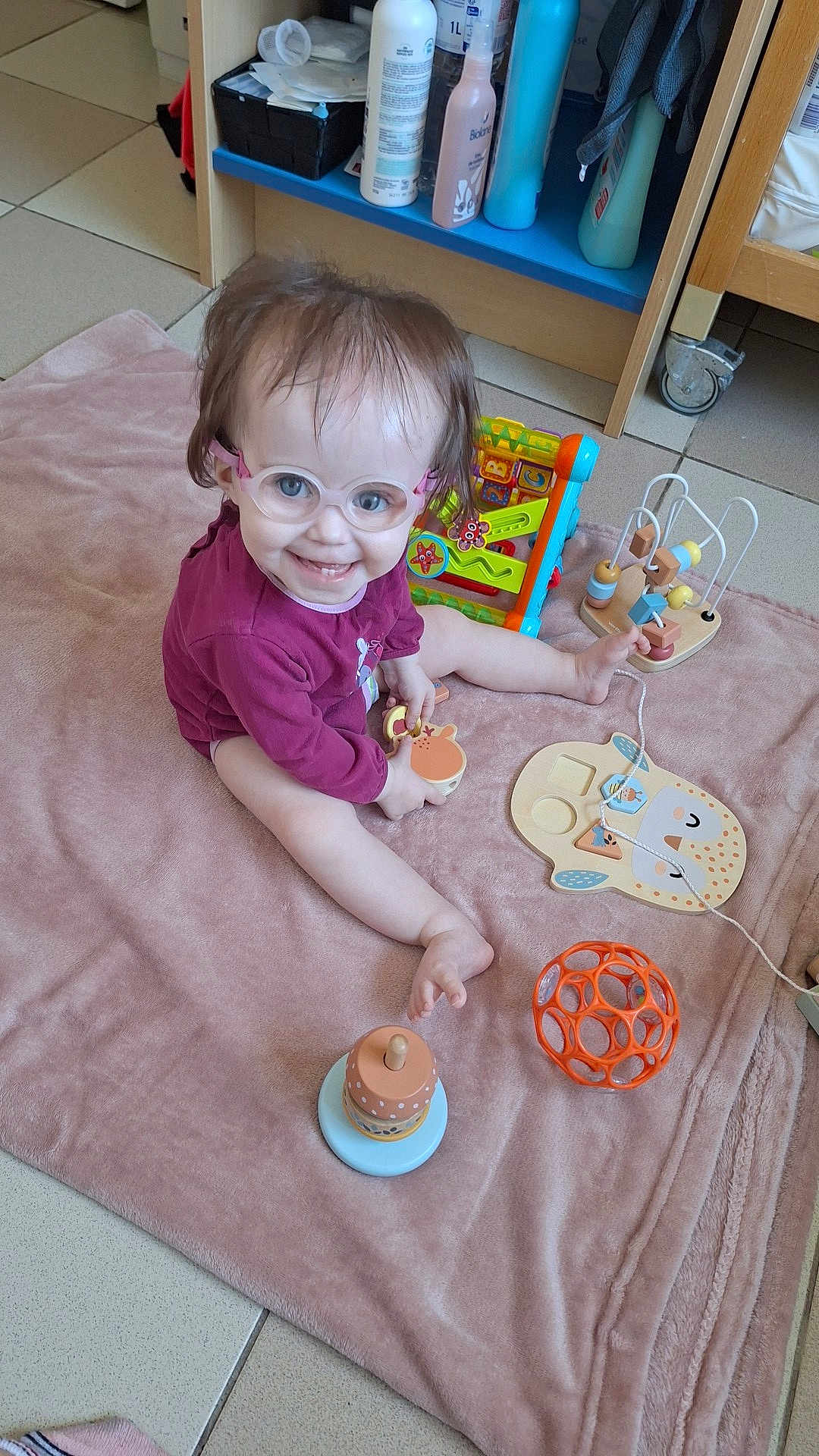 Lyna a rejoint le concours — aidez-le/la à gagner de superbes lots ! toddler, child, glasses, smile, toy, blanket, indoor, floor, sitting, happy, playtime, person, baby, wood, furniture, colorful, plastic, head, face, clothing