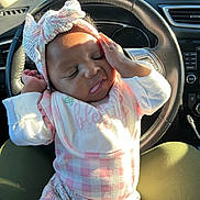 Ka Leyah is registered to the contest to win money with this photo: baby, infant, child, headband, bow, bib, clothing, car_interior, steering_wheel, sunlight, seat, lap, sleepy, cute, portrait, person, hand, pink, pattern, relaxing