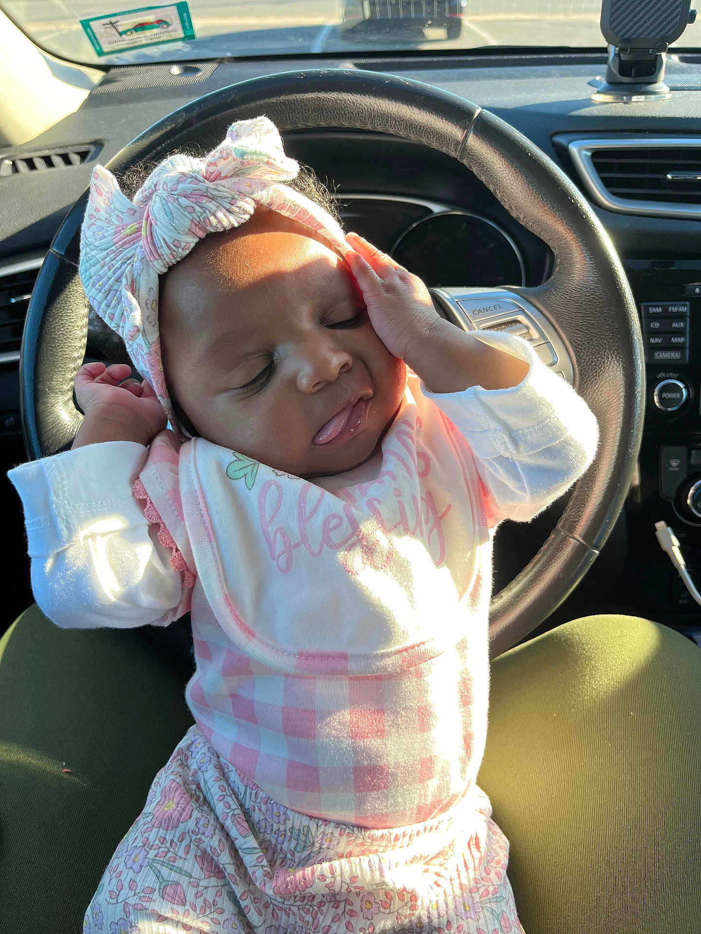 Ka Leyah is registered to the contest to win money with this photo: baby, infant, child, headband, bow, bib, clothing, car_interior, steering_wheel, sunlight, seat, lap, sleepy, cute, portrait, person, hand, pink, pattern, relaxing