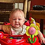 baby, child, walker, toy, red, floral_bib, indoor, wooden_chair, table, colorful, face, smiling, person, hands, playtime, cute, furniture, car_toy, expression, home