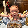 baby, child, indoor, activity_center, toy, surprised_expression, face, person, wooden_furniture, stroller, clothing, colorful, playtime, seat, happy, infant, cute, smiling, curly_hair, home