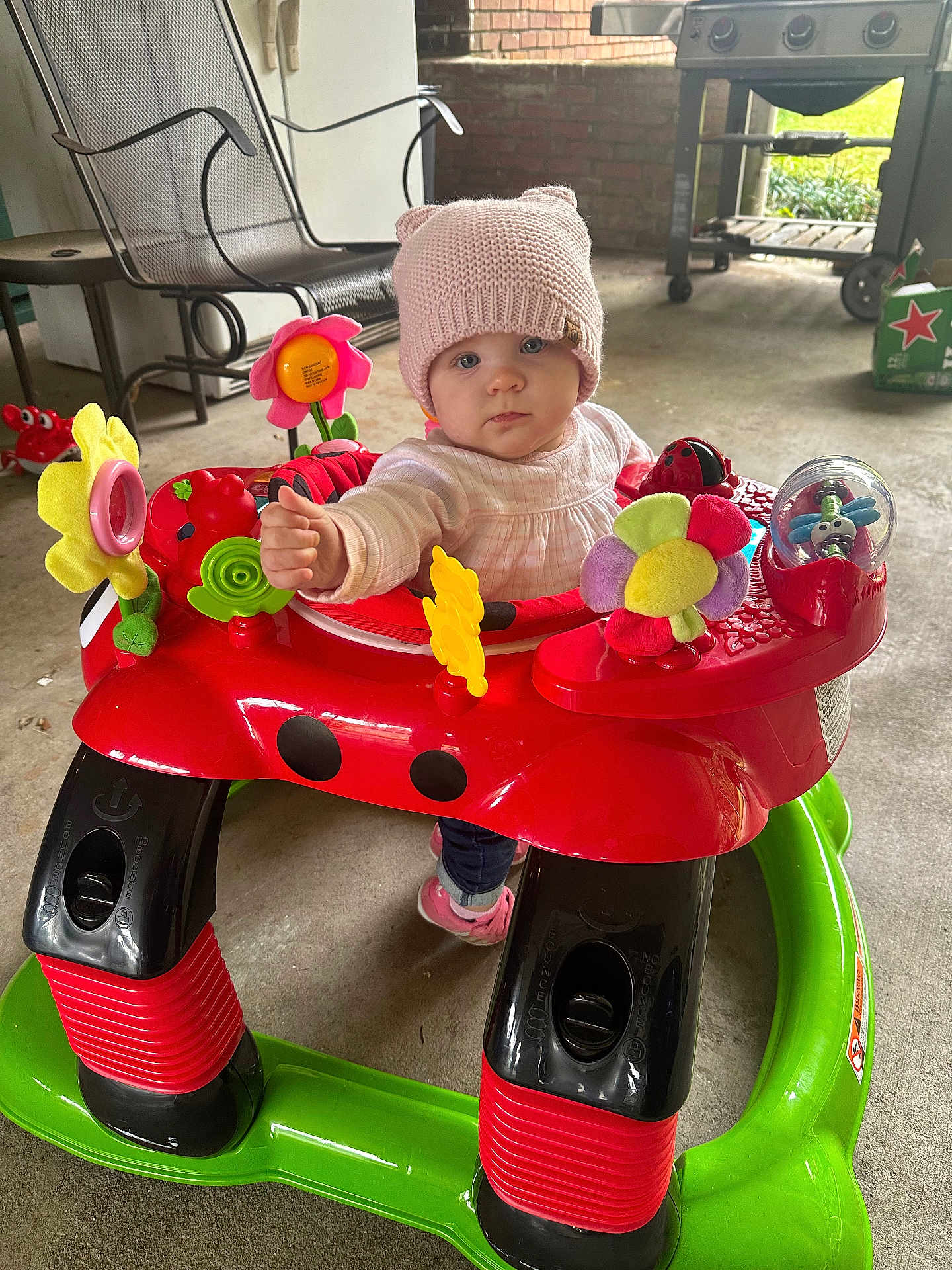 Layla joined the competition — help win amazing prizes! baby, child, walker, toy, pink_hat, flower_toy, indoor, porch, concrete_floor, curious, seated, colorful, playful, person, infant, cute, warm_clothing, footwear, smiling, face