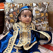 Adam a rejoint le concours — aidez-le/la à gagner de superbes lots ! baby, infant, child, traditional_clothing, blue_outfit, gold_embroidery, hood, velvet, pillow, couch, sitting, portrait, big_eyes, indoor, home_interior, textured_wall, fabric, ornate, decorative, cute